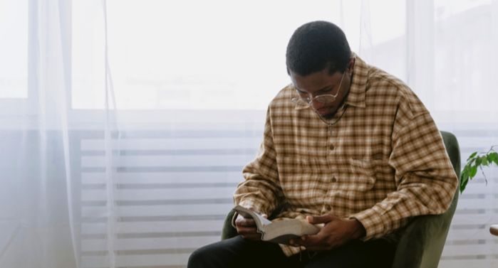 a brown-skinned Black man reading a book in a chair