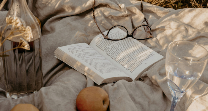 an open book on a picnic blanket with a pair of eyeglasses, some fruit, and a glass bottle and goblet