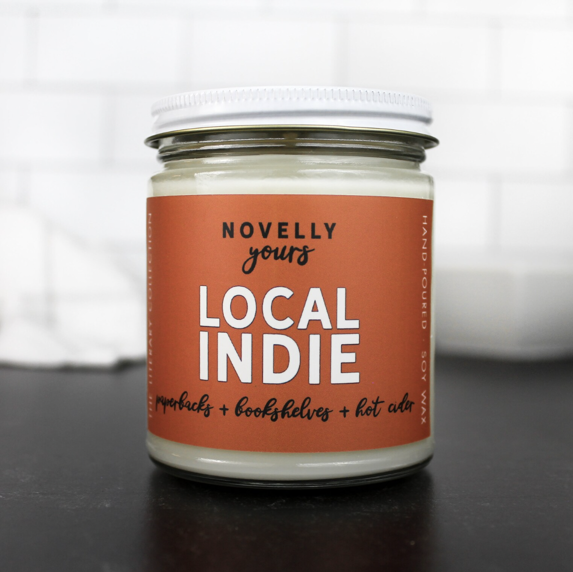 Image of a soy candle in a lidded glass jar with an orange label saying it's "local indie" scented