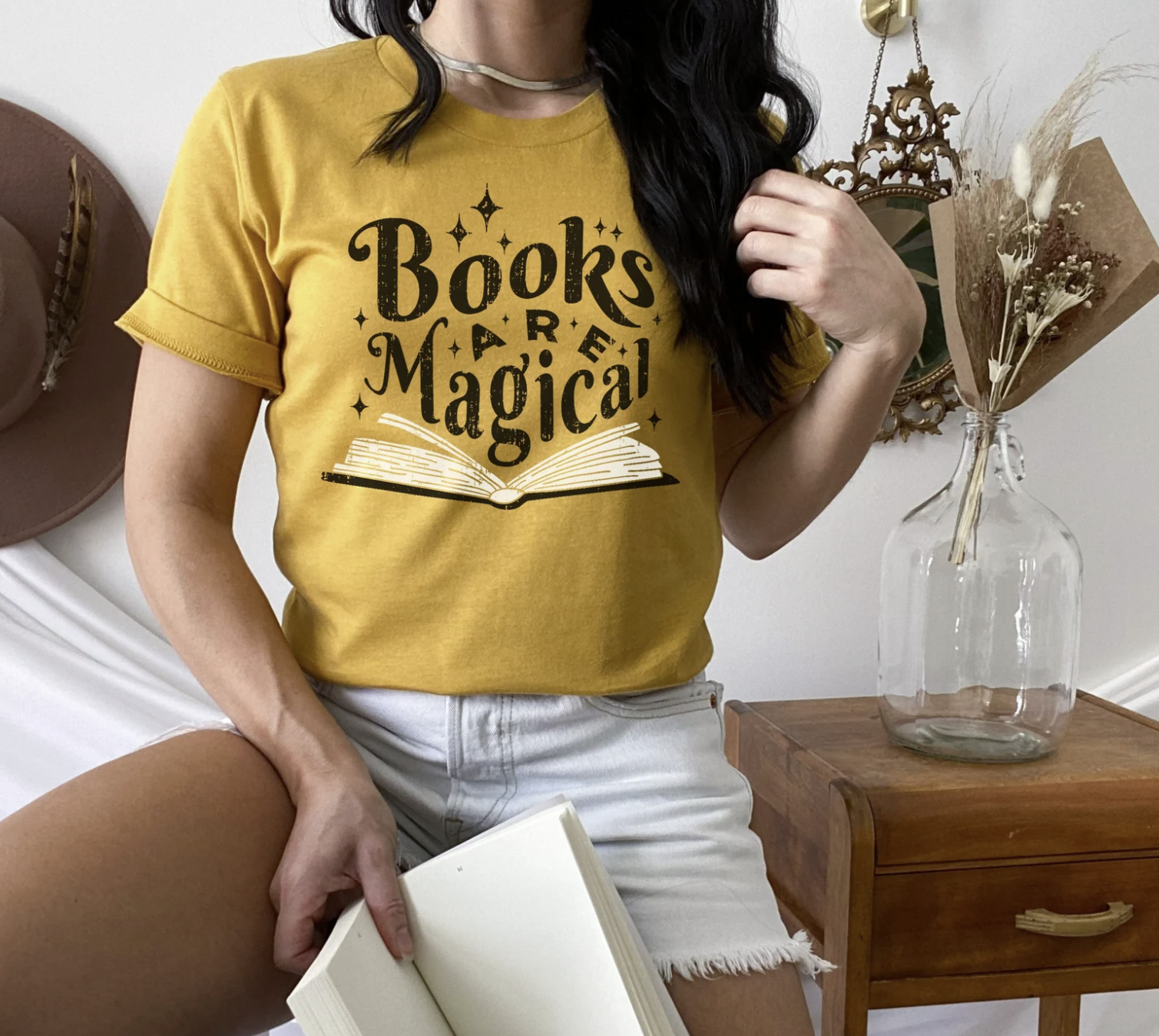 Picture of a white woman with dark hair posing with an open book in one hand and wearing a golden yellow shirt with an image of an open book with the words "books are magical" coming out of it