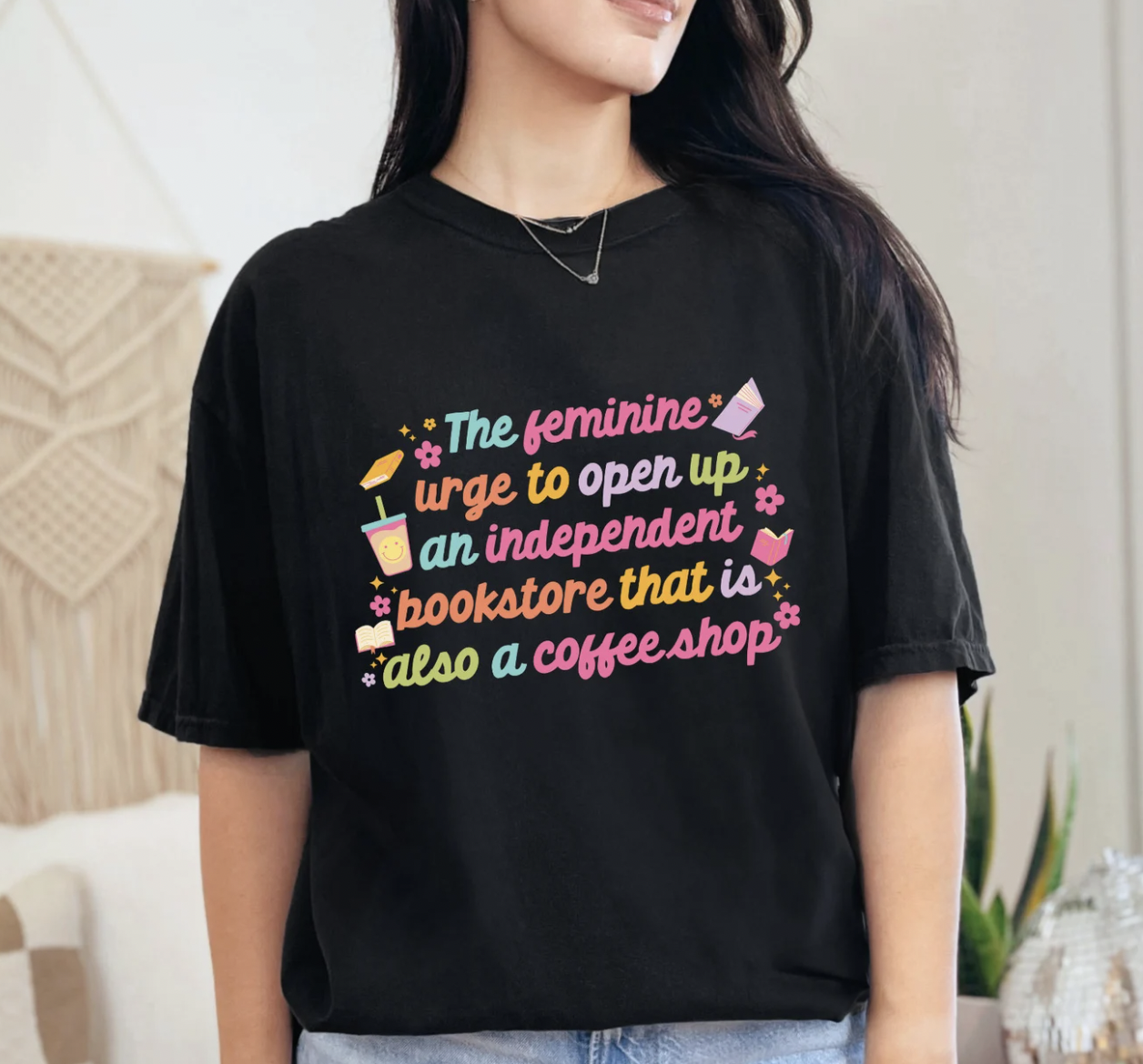 Image of a person wearing a black t-shirt with colorful screen printing that says "the feminine urge to open an independent bookstore that is also a coffee shop"