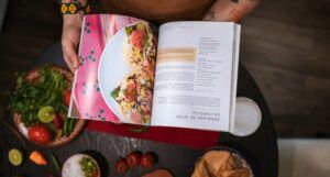 a medium light skinned person with tattoes holding open a cookbook in a kitchen