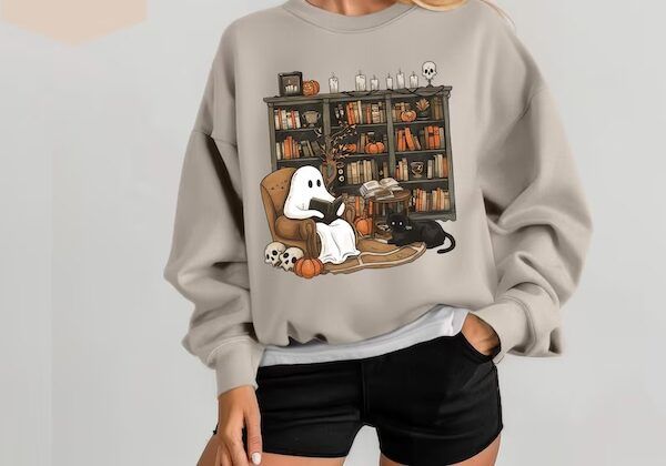 light grey crewneck sweater with a design on the front of a ghost reading a book in an armchair in front of a wall of bookshelves containing books, candles, and pumpkins/ There is a black cat at the ghost's feet