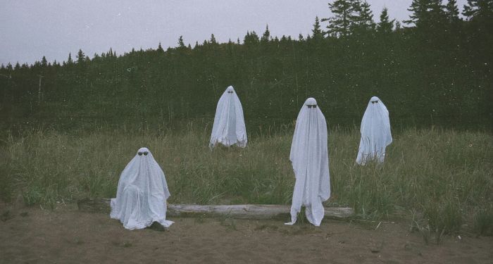 four people dressed in white sheets with black sunglasses to appear like ghosts in a field of grass and sand