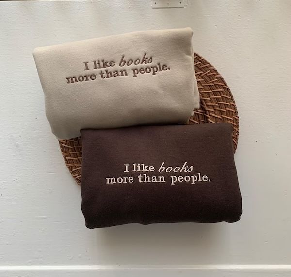 two folded crewneck sweaters, one tan and one dark brown. Both have embroidered text on the front that reads "I like books more than people"