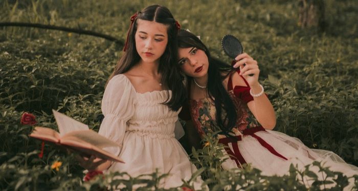 two fair-skinned women dressed in a medieval:fantastical theme sitting in a field,;one is reading a book