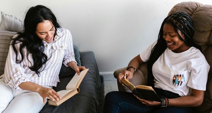 two women, one with light tan skin and the other with brown skin, reading books and laughing with each other