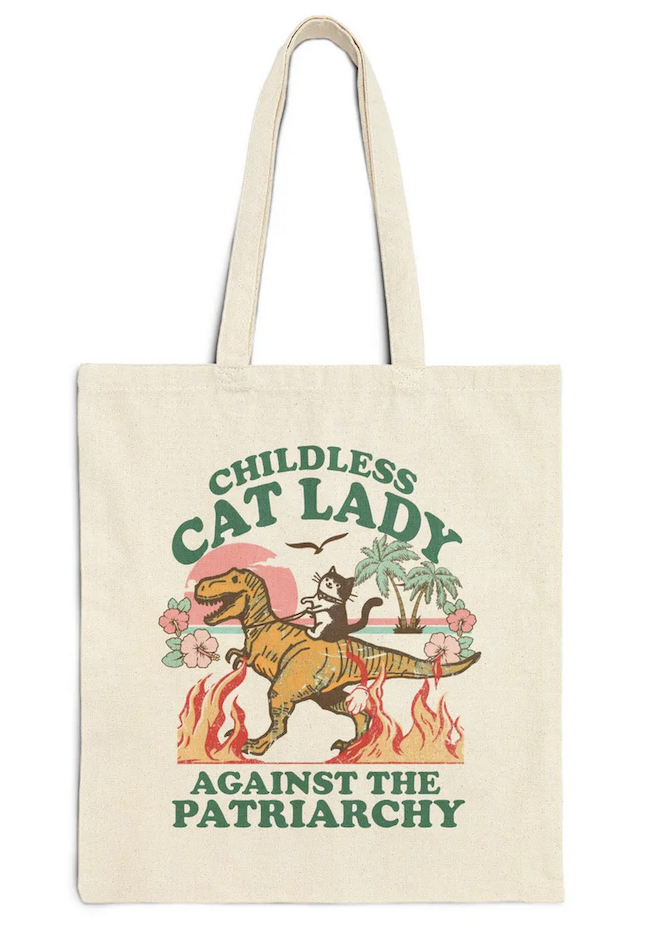 a tote back with an illustration of a cat riding a dinosaur that says "childless cat lady against the patriarchy"