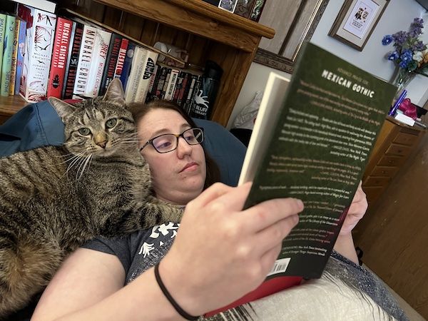 a brown tabby cat curled up near a woman's head while she reads in bed