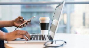 woman holding phone with hands on laptop keyboard in an office with a to go coffee on eyeglasses on desk