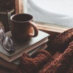 a coffee mug resting on a pile of books next to a blanket by a window with snow outside