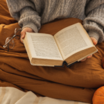 person wearing a sweater and reading in bed with decorative pumpkins around