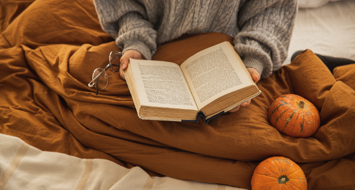 person wearing a sweater and reading in bed with decorative pumpkins around