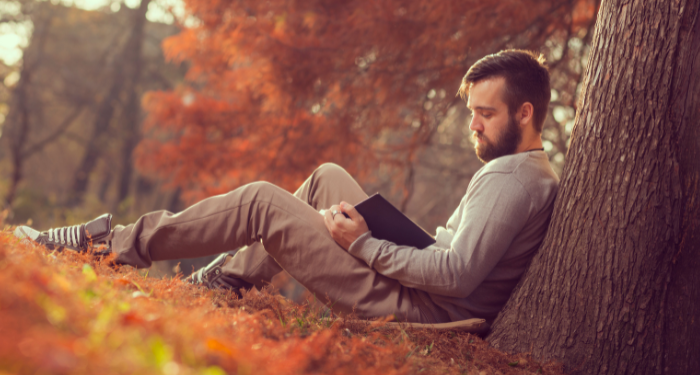 a man with light skin reads outside, surrounded by autumn leaves