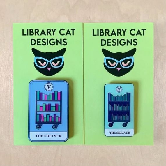 image of a green cardstock card that says Library Cat Designs displaying a blue tarot card with a cart of books that days The Shelver 