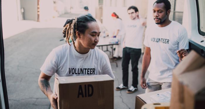 people volunteering for relief causes. Two brown-skinned men and a white man are wearing white t-shirts that say "volunteer" on them and are moving cardboard boxes labeled "aid"