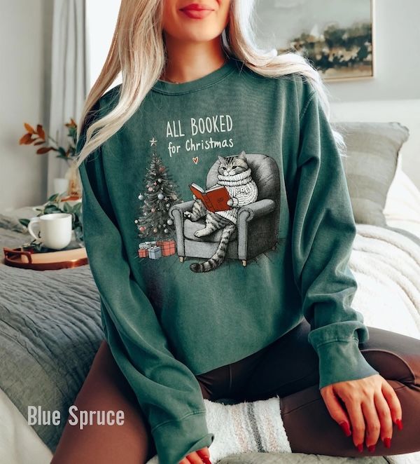 a white woman wearing a heathered green crewneck sweater with a graphic of a cat wearing a knit sweater sitting in an armchair reading a book next to a Christmas tree