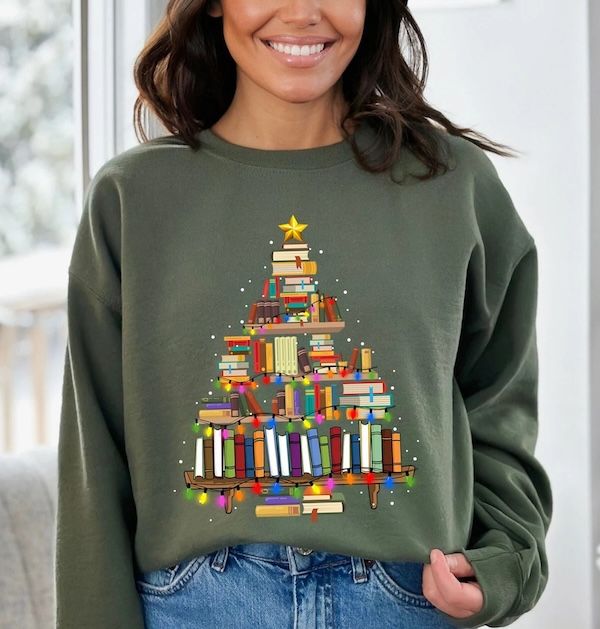 a woman with tan skin wearing an olive green crewneck sweater with a graphic of books on bookshelves arranged to look like a Christmas tree