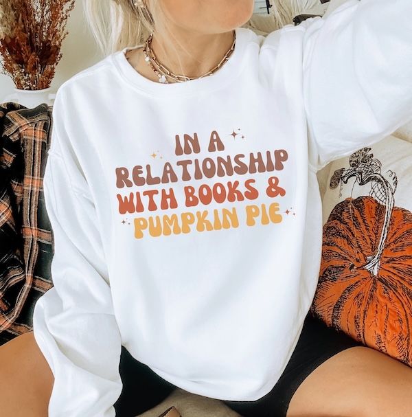 a white woman wearing a white crewneck sweatshirt with text that reds "in a relationship with books & pumpkin pie"