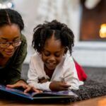 A Black woman and child reading a picture book on the floor of a living room with a Christmas tree and fireplace in the background