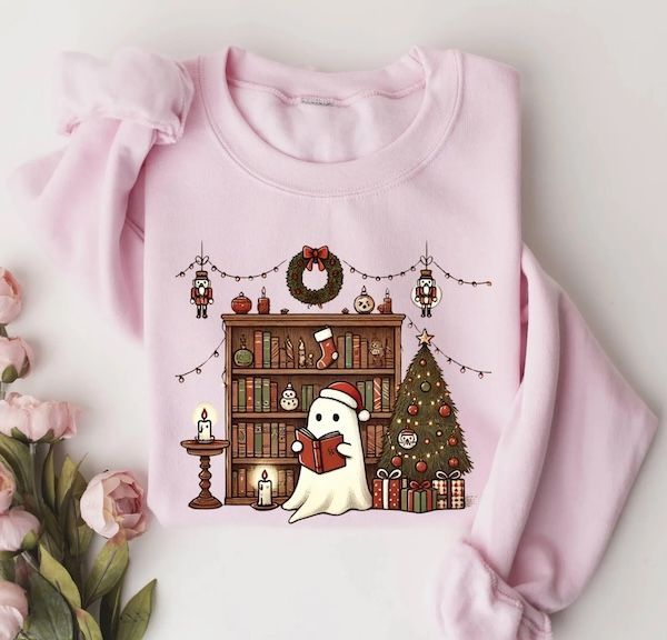 a light pink crewneck sweater with a graphic of a small white ghost wearing a Santa hat and reading a book in front of a bookshelf with holiday decorations and a Christmas tree