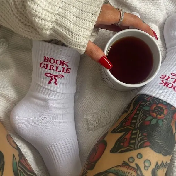 a hand with red nails mug of something near crossed, tattooed legs. the free are wearing white crew length socks with read letters that say "book girlie' with a red bow