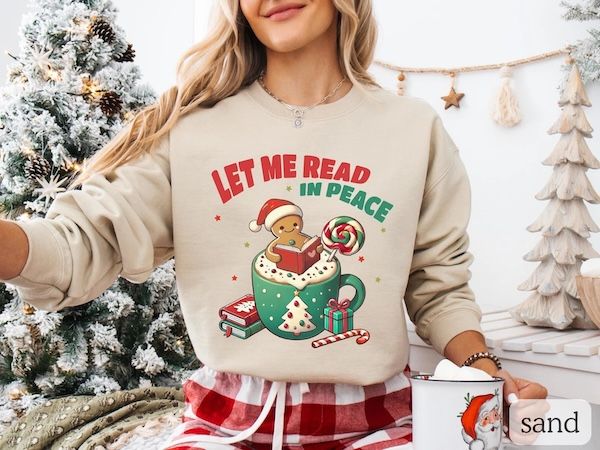 a white woman wearing a tan crewneck sweater with text that reads "Let Me Read in Peace" hovering over a graphic of a gingerbread man sitting inside a coffee mug reading a book