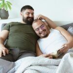 two men reading in bed and smiling, one with his arm around the other