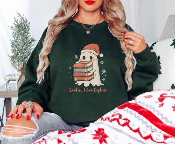 a white woman in a dark green sweater with a graphic on the front of a small ghost wearing a Santa hat and holding a small pile of books. Text beneath the ghost reads "Santa, I can explain"