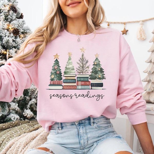 a white woman wearing a pink crewneck sweater with cursive text that reads "season's readings" beneath a graphic of four assorted style of Christmas trees atop piles of books