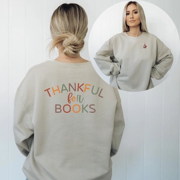 a white woman wearing a tan crewneck sweater. the. image of the front shows a small turkey reading a book in the upper left corner of the sweater. Image of the back shows text that reads "thankful for books"