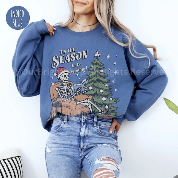 a white woman wearing a blue crewneck sweater with text that reads "'Tis the Season to be Reading" hovering over a graphic of a skeleton wearing a Santa hat reading by a Christmas tree