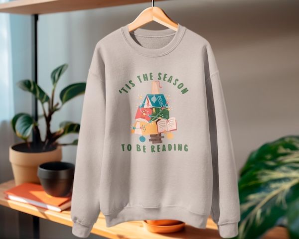 a grey crewneck sweater on a hanger with text that reads "'tis the season to be reading" bordering an image of a small christmas tree make of books