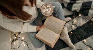 fair skinned, red-haired person sitting cross-legged and reading with a cup of hot chocolate