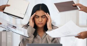 brown-skinned woman with her hands at her temples and eyes closed like she has a headache. She is stting in front of a laptop and people in all directions are handing her things, like reports, documents, and a tablet