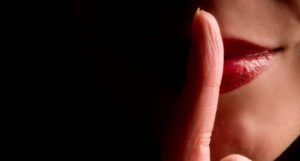 Image of a finger in front of a mouth making a gesture about a secret