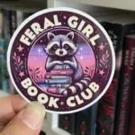 a sticker of a raccoon on a stack of books with the text Feral Girl Book Club