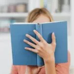 fair-skinned person holding a light blu book in front of their face