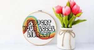 image of a colorful cross stithc that says "support your local library" beside a vase of tulips