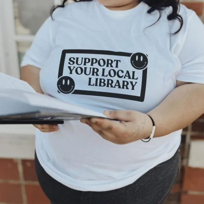 a white t-shirt with a simple, black, retro-inspired design including the words "support your local library" and smiley faces 