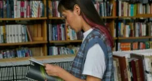 a tan skinned Asian woman with a red streak in her hair reads a book in a library