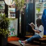 woman sitting in front of bookstore with big windows reading a book